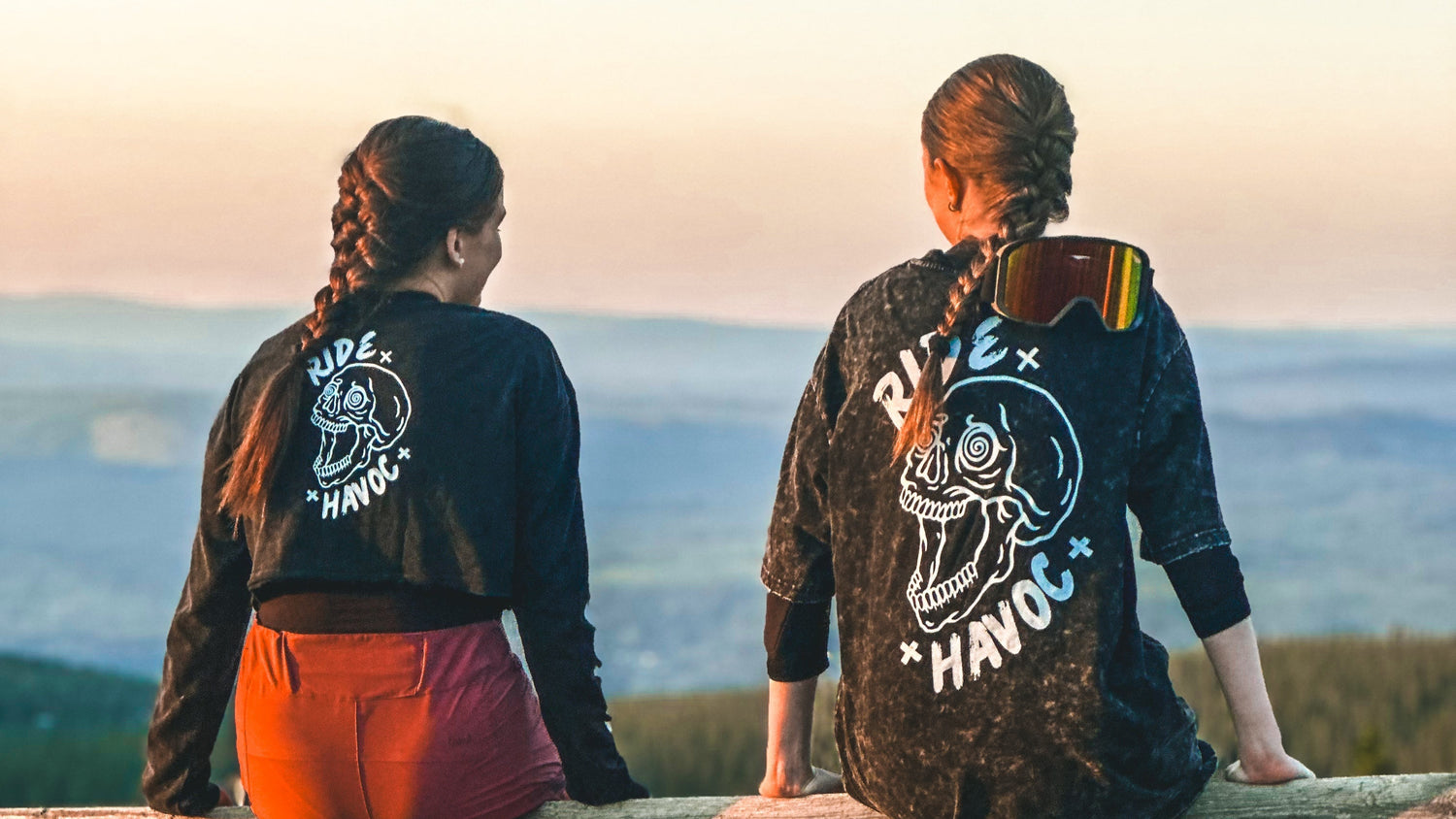 Two people sitting on a wooden fence with bicycles at sunset, wearing Ride With Havoc black tees with visible skull branding.